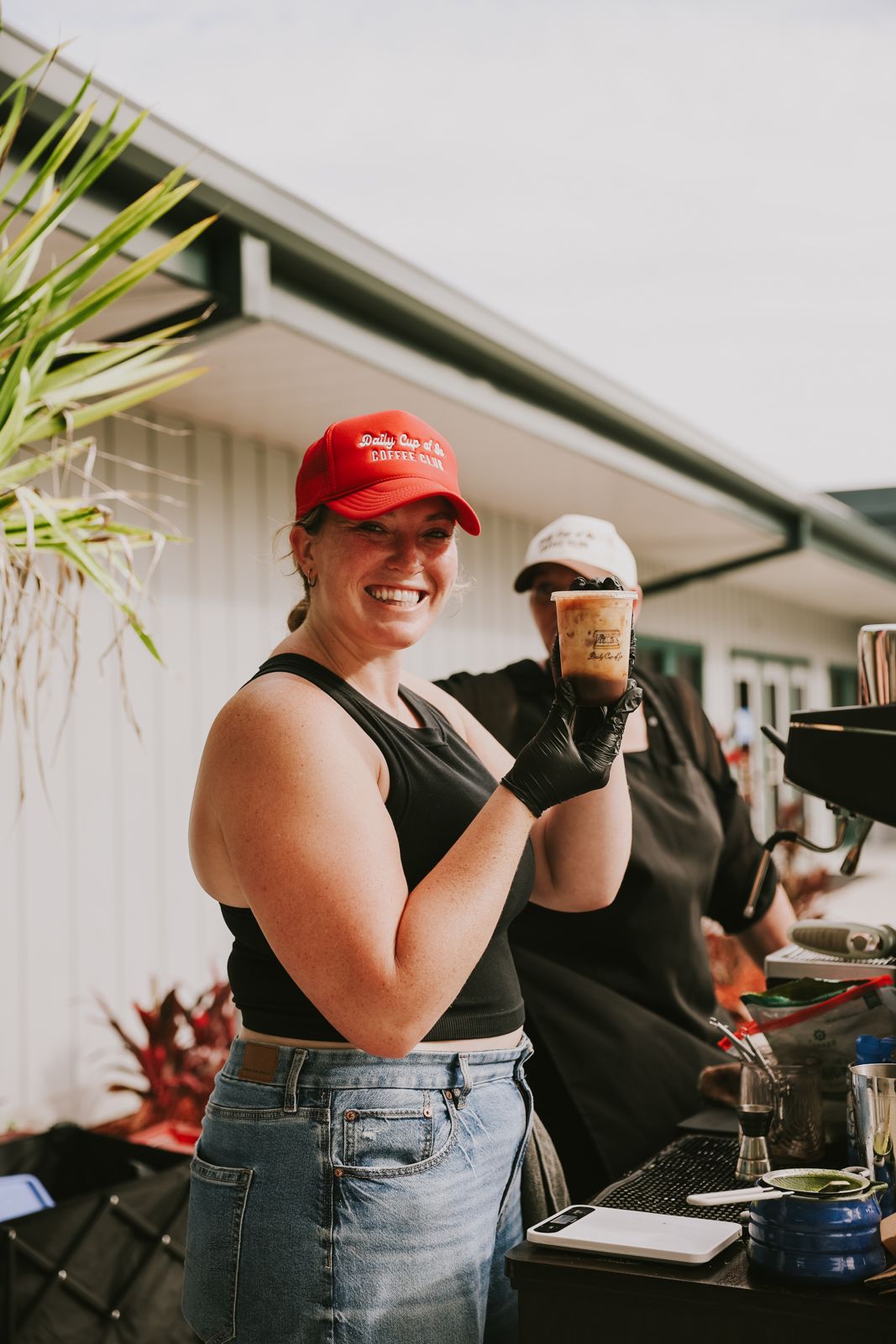 Barista in a red Daily Cup of Jo cap holding up an iced latte at the cart